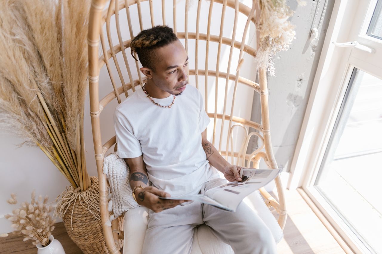 Man with tattoos reads a magazine in a bright, cozy setting with natural decor.